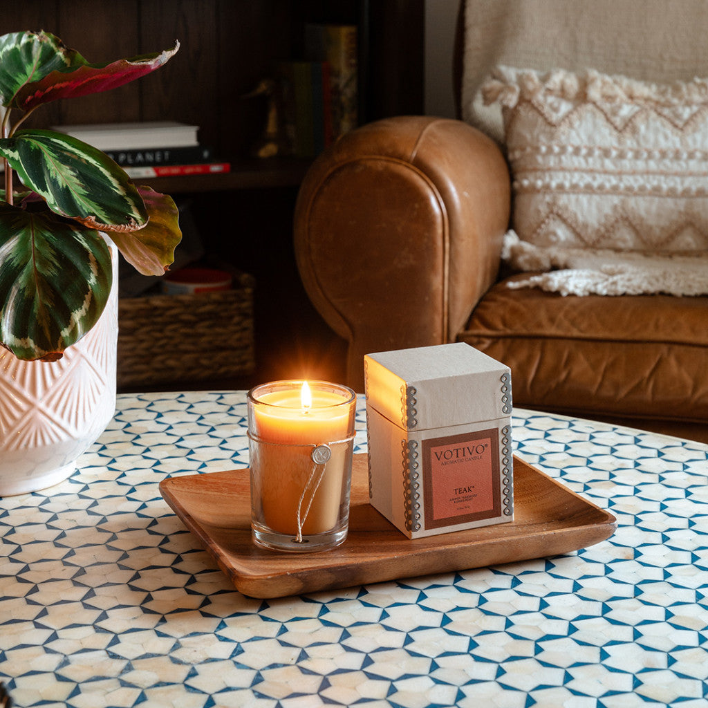A lit Votivo Teak 6.8 oz. Aromatic Candle in a glass jar and its box rest on a wooden tray atop a patterned table, with a potted plant and brown leather armchair in the background.