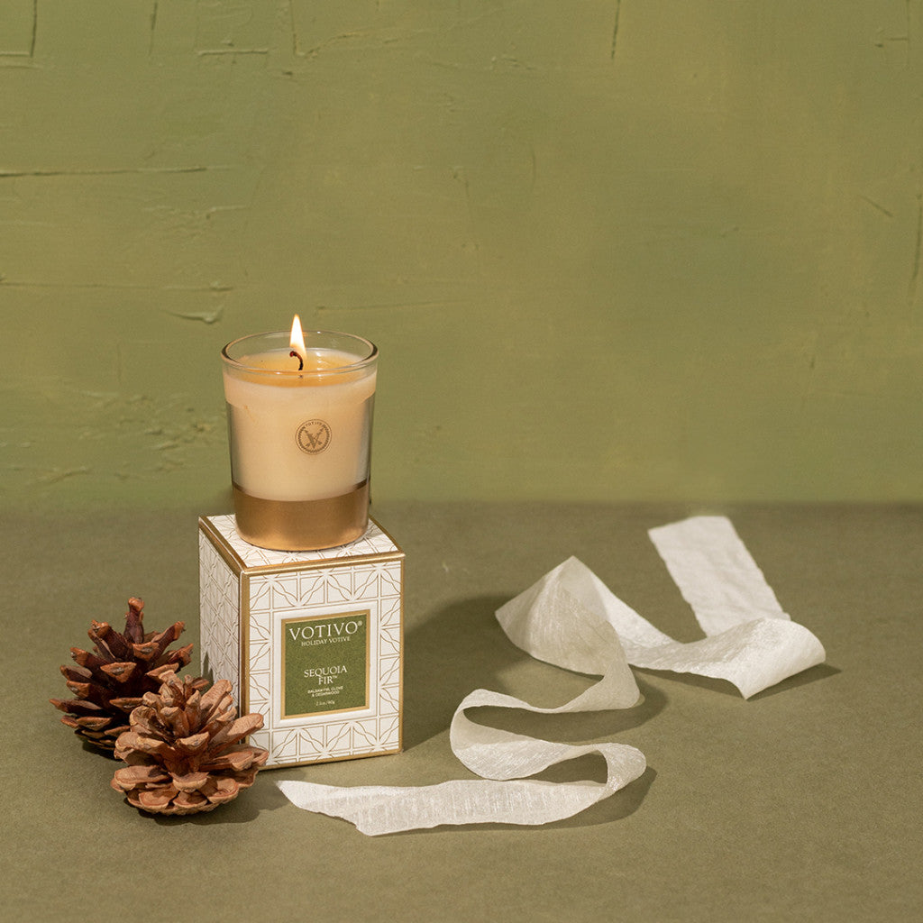 The Votivo Sequoia Fir Holiday Votive candle rests atop its box beside two pine cones and white ribbon on a green textured background—ideal for adding the fresh scent of sequoia fir to your festive décor.