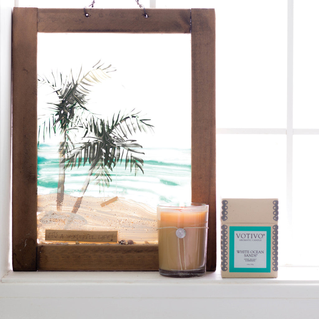 A framed palm tree painting sits on a windowsill next to a tan candle in a glass jar and a Votivo White Ocean Sands 6.8 oz. Aromatic Candle box, as sunlight streams through the window behind them.