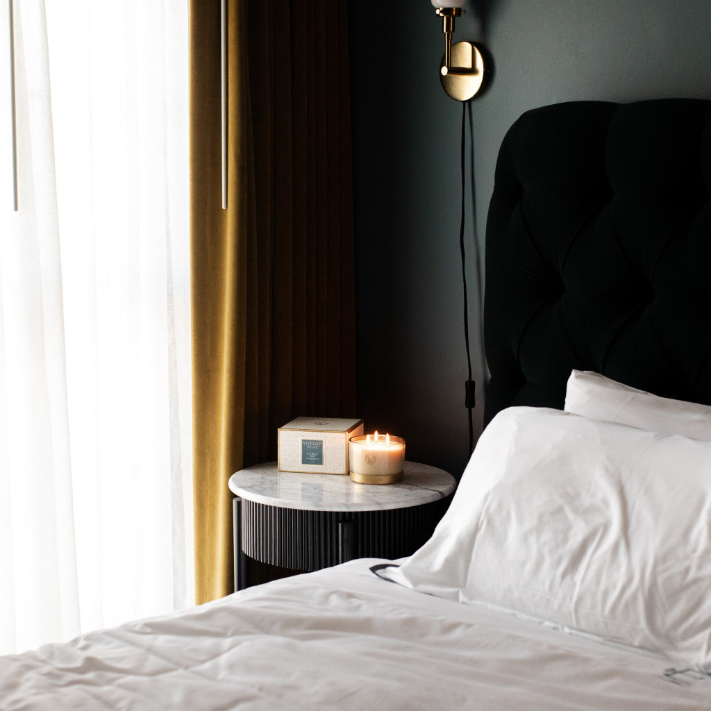 A neatly made bed with white linens sits by a round marble nightstand holding a lit Votivo Icy Blue Pine Holiday 3 Wick Candle and a small box. Soft natural light through sheer curtains creates a cozy, relaxing atmosphere.