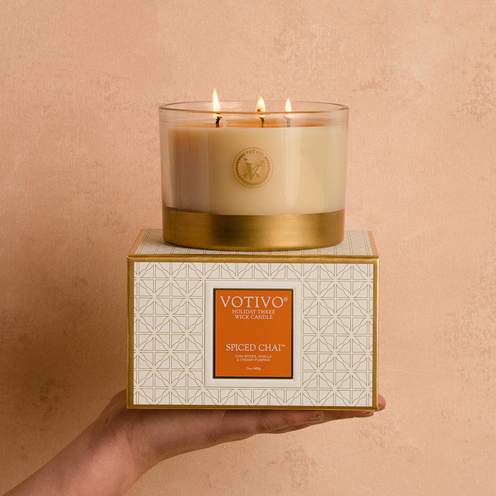 A hand holds a Votivo Spiced Chai Holiday 3 Wick Candle box with a geometric pattern, topped by the lit soy blend wax candle in a clear glass vessel with a gold base. The background is warm beige.