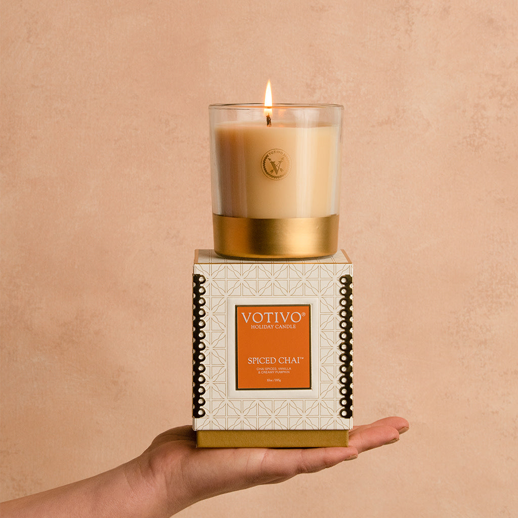 A hand holds a Votivo Spiced Chai 10 oz. Holiday Candle made from soy blend wax in a glass container, with the box beneath, set against a neutral beige background.