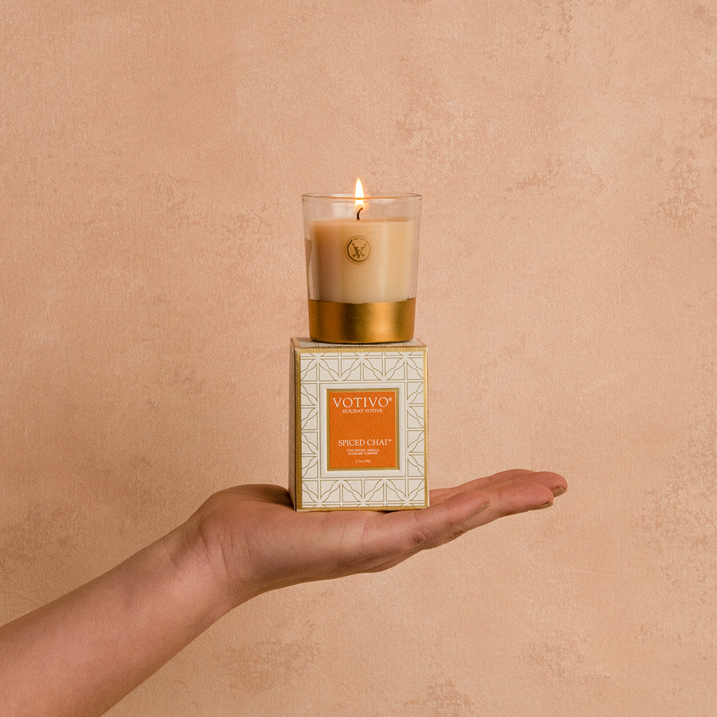 A hand holds a lit Votivo Spiced Chai Holiday Votive made with soy blend wax in a glass container, resting on its decorative box against a beige textured background.