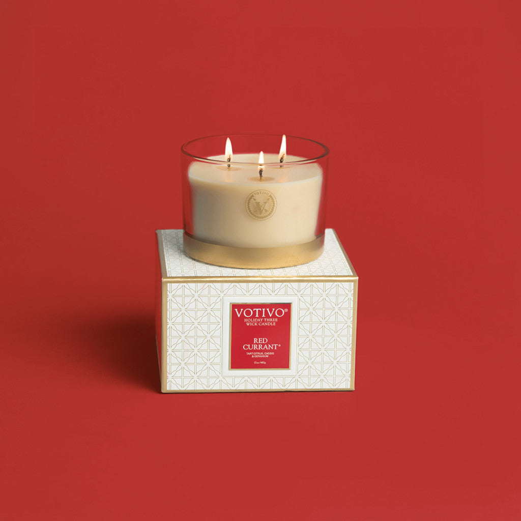 The Votivo Red Currant Holiday 3 Wick Candle sits in a clear glass jar on its white and gold patterned box, displayed against a solid red background.
