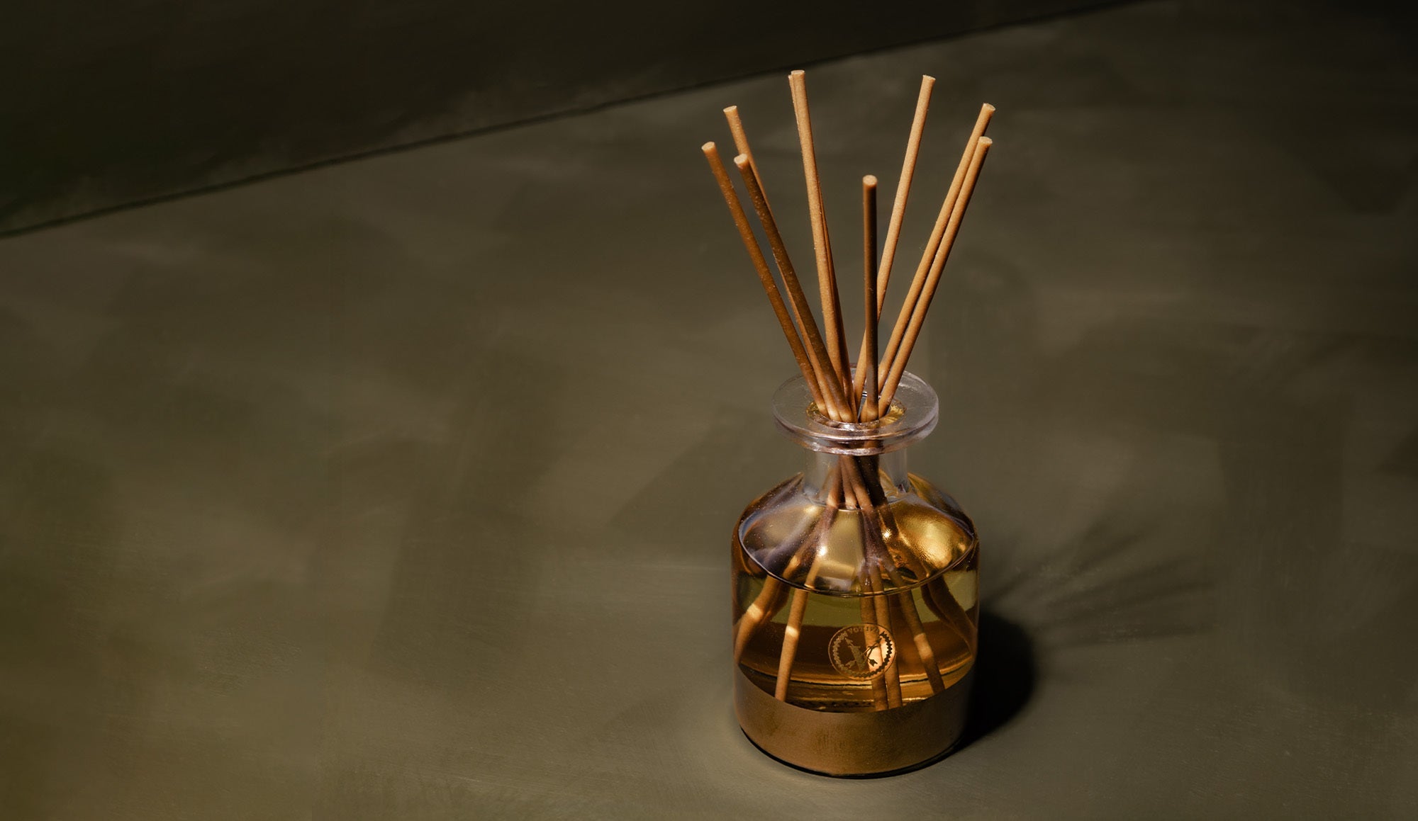A gold-colored glass reed diffuser with several wooden sticks is placed on a smooth, dark green surface under soft lighting.