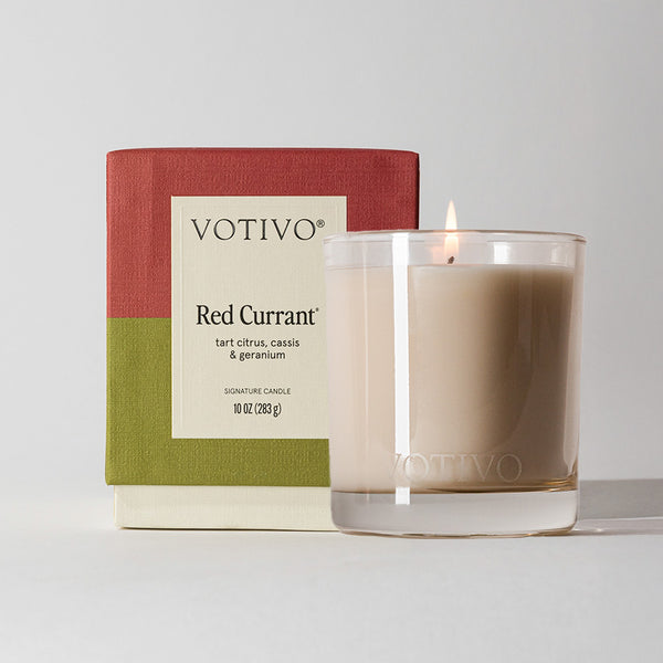A lit Votivo Red Currant 10 oz. Signature Candle, crafted with soy blend wax, sits in a clear glass holder beside its red and green box labeled Red Currant and scent notes: tart citrus, cassis & geranium.