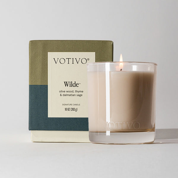 A lit Votivo Wilde 10 oz. Signature Candle in a clear glass jar sits before its green, tan, and blue box. Featuring soy blend wax, it offers a unique olive wood, thyme & dalmatian sage fragrance.