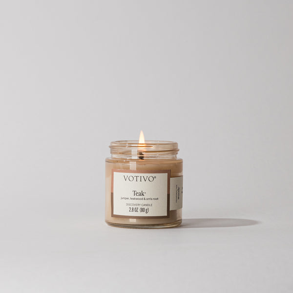 A small Votivo Teak Discovery Candle glows with a gentle flame, filling the area with a warm, woody scent. The candle sits against a plain light gray background.