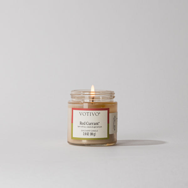 A Votivo Red Currant Discovery Candle glows softly, its soy blend wax jar resting on a plain white surface with a light gray background.