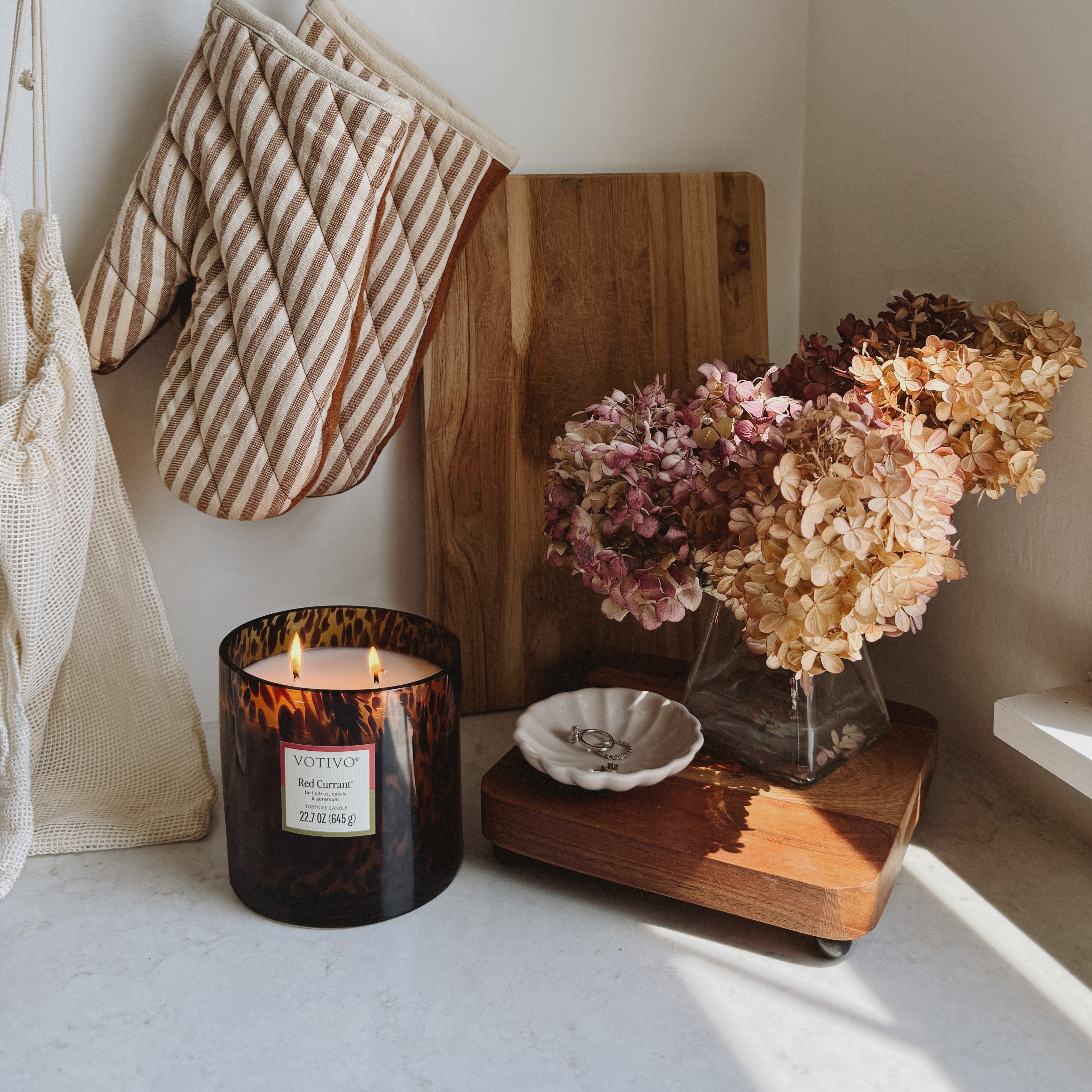 A Votivo Red Currant Tortoise Candle, dried hydrangea flowers in a glass vase, a ring dish, striped oven mitts, and a wooden cutting board are arranged on a bright kitchen countertop.