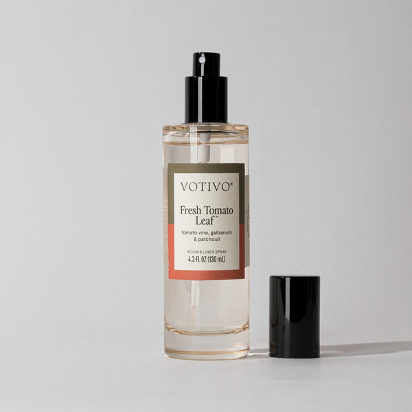The Votivo Fresh Tomato Leaf Room & Linen Spray stands upright on a light gray surface, featuring a clear bottle with black spray nozzle and cap beside it—ideal for adding fabric-safe freshness.