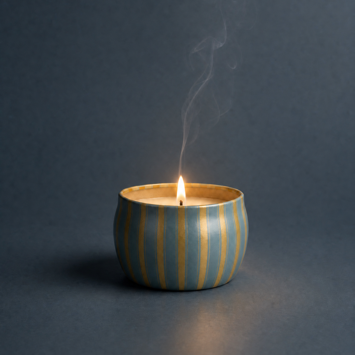A lit Icy Blue Pine Tin Candle with blue and gold striped packaging on a blue background