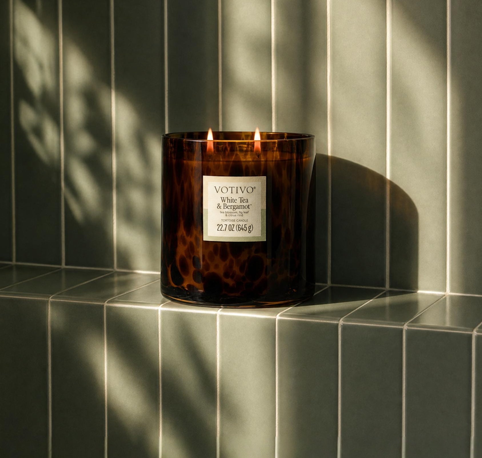 A Votivo White Tea & Bergamot Tortoise Candle with two wicks rests on a tiled ledge, its brown glass casting soft shadows in the warm sunlight. Made from a soy blend wax, it releases a delicate fragrance. Behind it, light green vertical tiles form the backdrop.