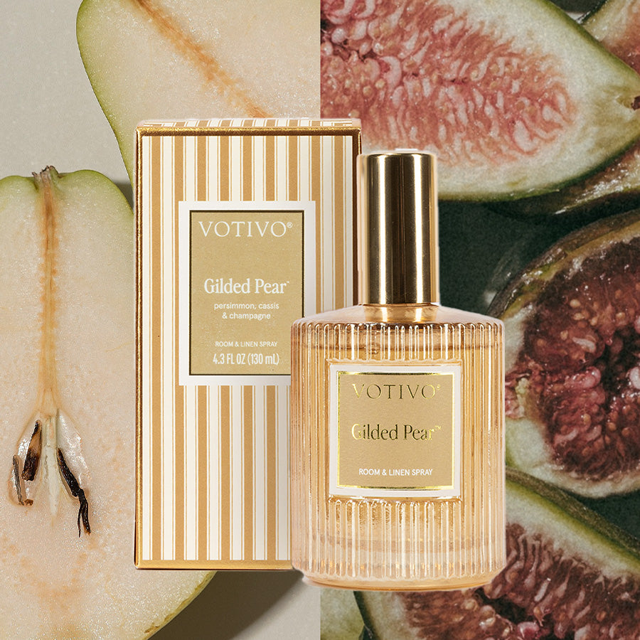 A Votivo Gilded Pear Room & Linen Spray bottle and striped box are displayed against a backdrop of close-up pear and fig slices.