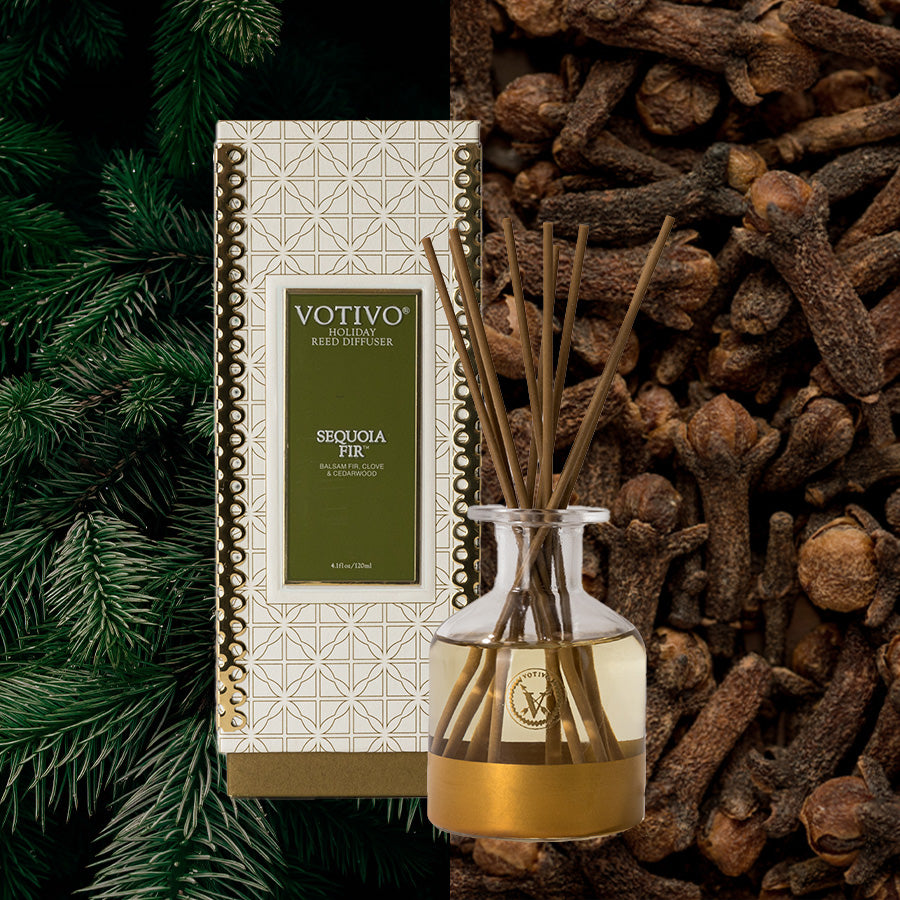 The Votivo Sequoia Fir Holiday Reed Diffuser with box is displayed, surrounded by fir branches and whole cloves, with rattan reeds highlighting its festive, natural fragrance.