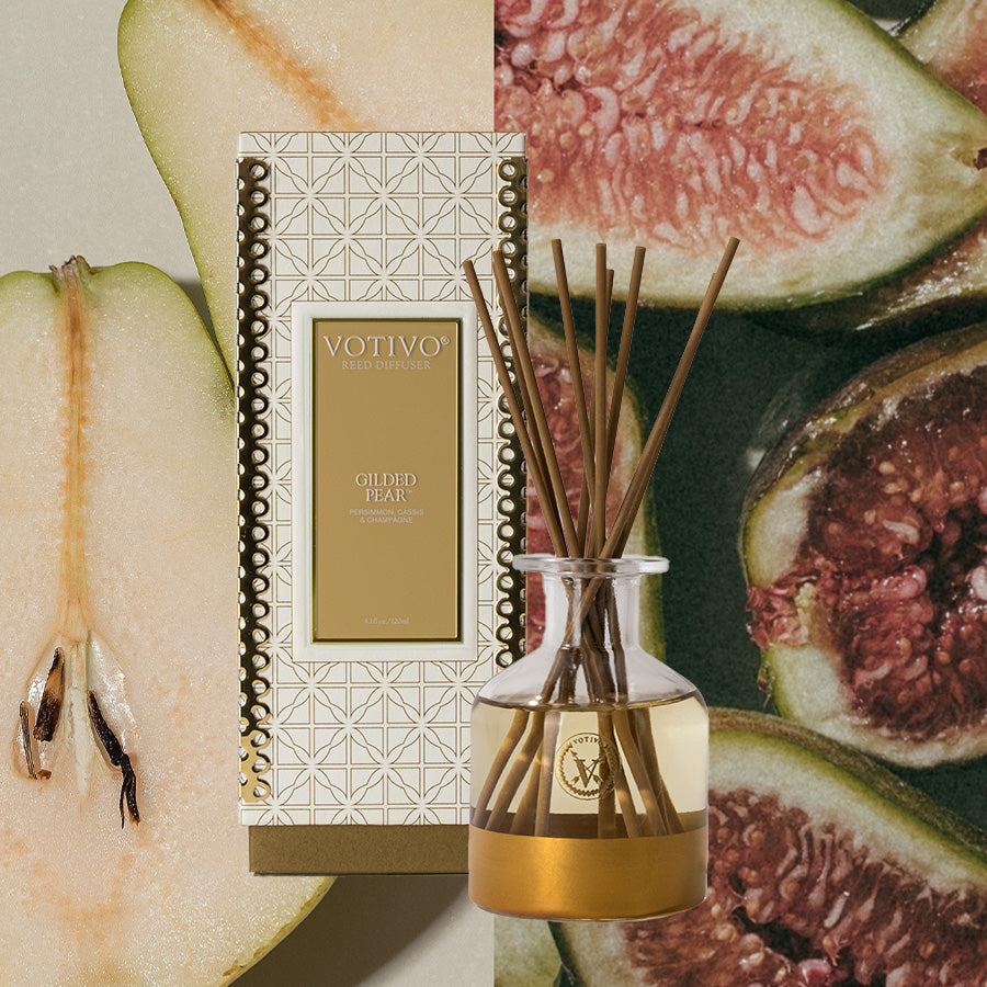 The Votivo Gilded Pear Holiday Reed Diffuser in gold packaging sits before a sliced pear and fresh figs, emphasizing its elegant and fruity fragrance.
