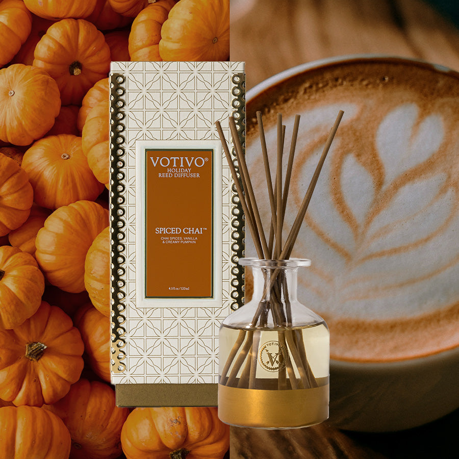 A Votivo Spiced Chai Holiday Reed Diffuser with its box and elegant rattan reeds is showcased against a close-up of pumpkins and a chai latte with latte art.