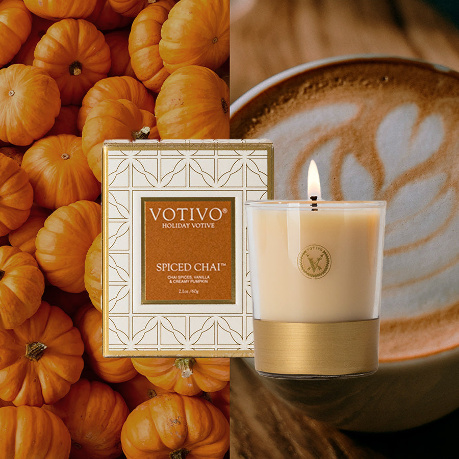 The Votivo Spiced Chai Holiday Votive is displayed in front of its box, flanked by small orange pumpkins on the left and a latte with leaf-shaped foam art on the right.