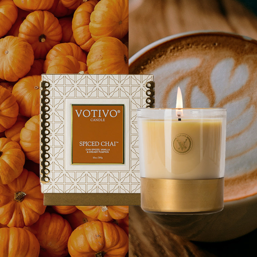 A Votivo Spiced Chai 10 oz. Holiday Candle sits before its festive box, surrounded by small pumpkins and a latte with heart-shaped foam.