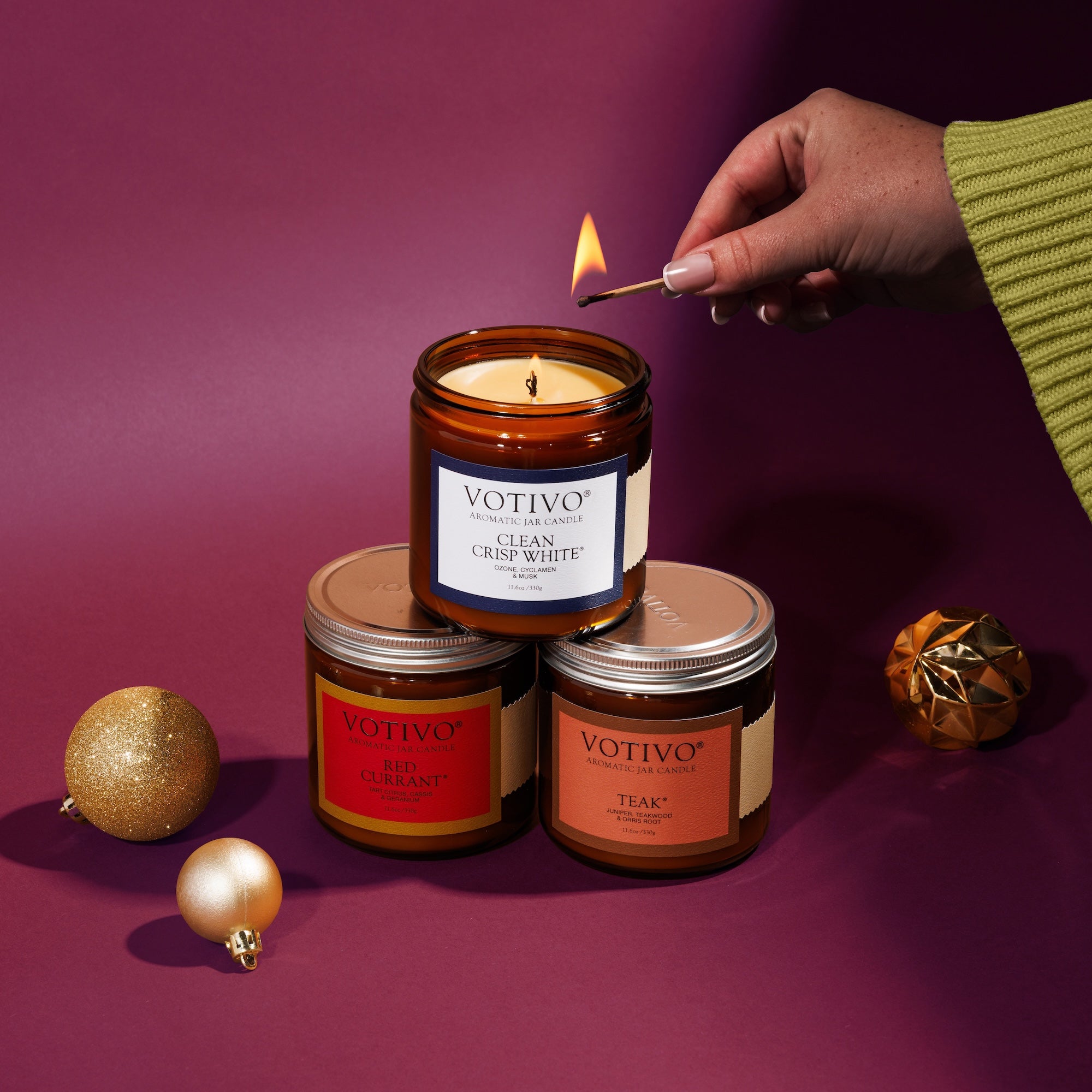 A hand in a green sleeve lights a Votivo Limited Edition Candle Set—Red Currant, Clean Crisp White, and Teak Large Amber Jars—arranged with gold ornaments on a purple background.