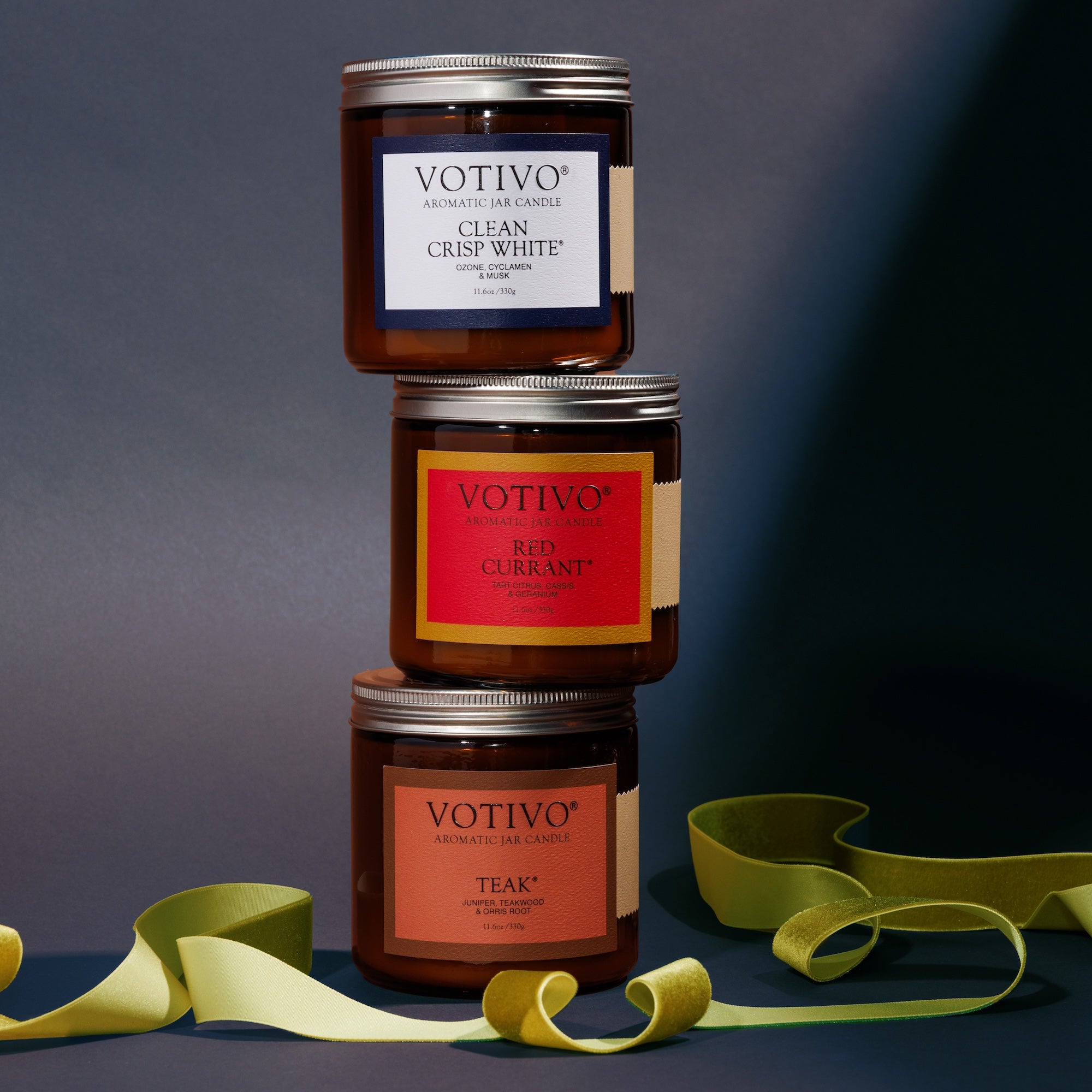 The Votivo Limited Edition Candle Set features large amber jars in Red Currant, Clean Crisp White, and Teak, elegantly stacked and tied with a yellow ribbon against a dark backdrop.