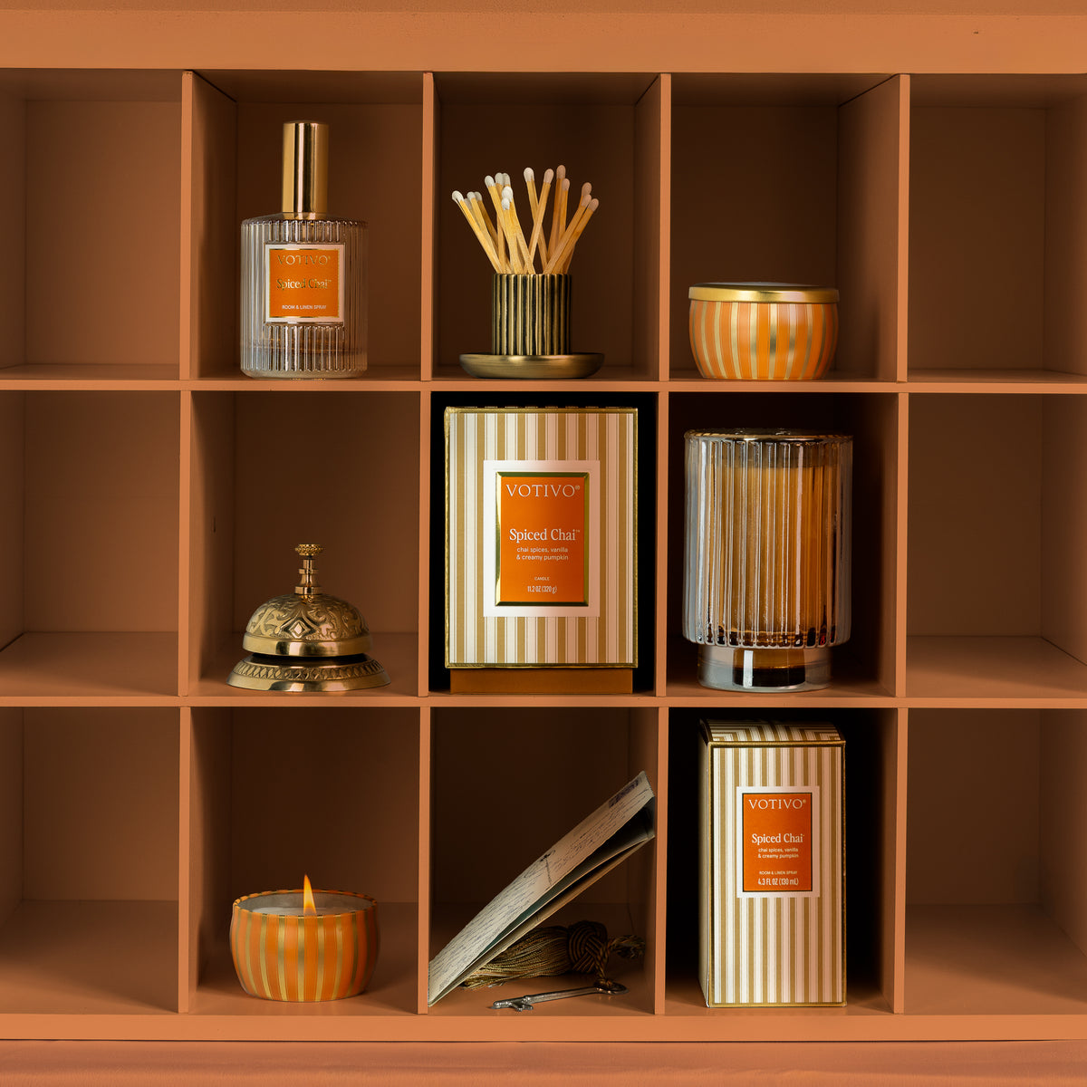 A warm-toned shelving unit with multiple cubbies displaying coordinated spice chai home fragrance and decor items, including reed diffusers, candles, and a boxed scented candle. The products feature striped packaging and orange labels.