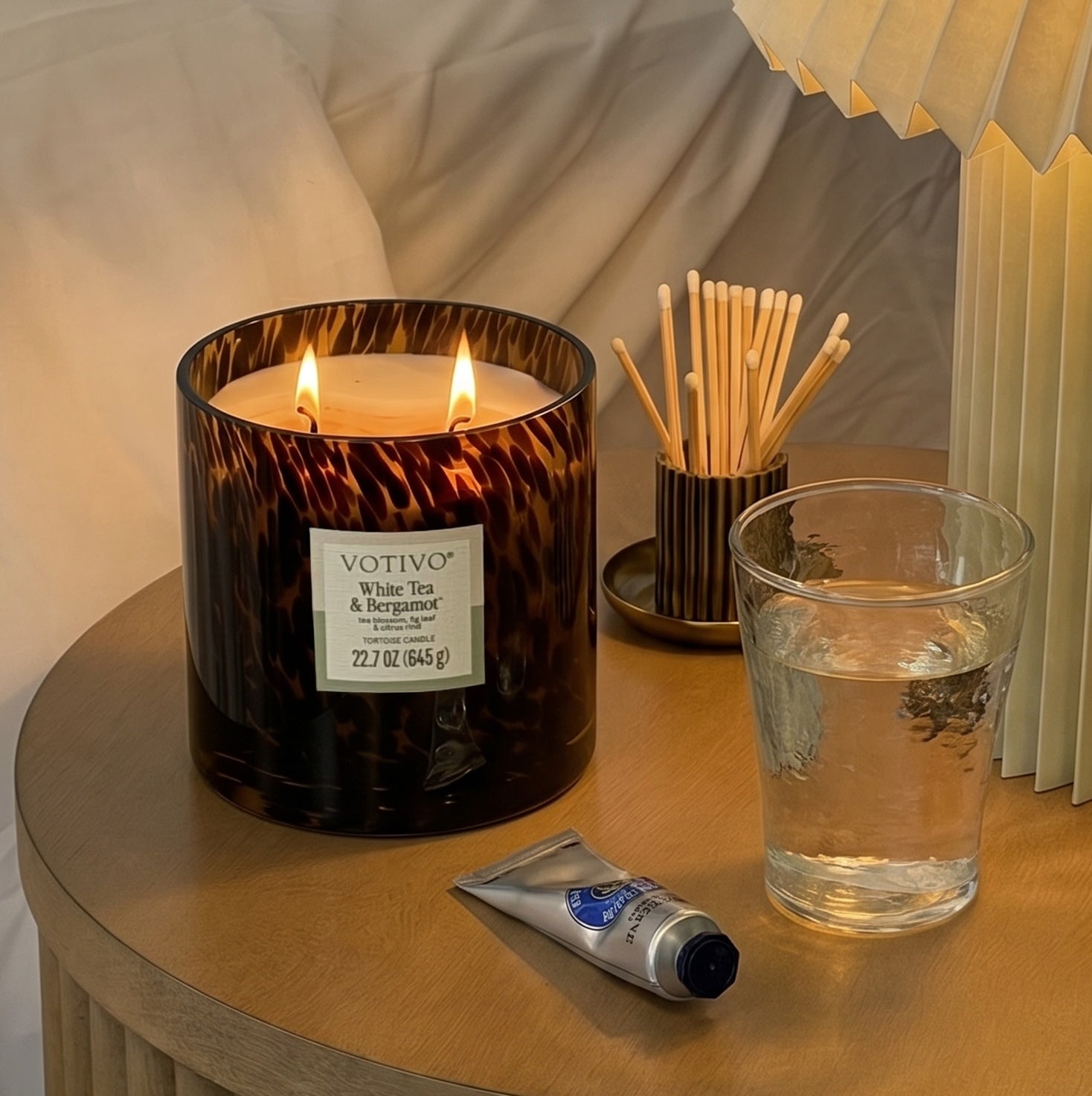 A lit Votivo White Tea & Bergamot Tortoise Candle sits on a wooden nightstand beside a glass of water, a small tube of hand cream, a lamp, and a tortoiseshell container filled with wooden matches.