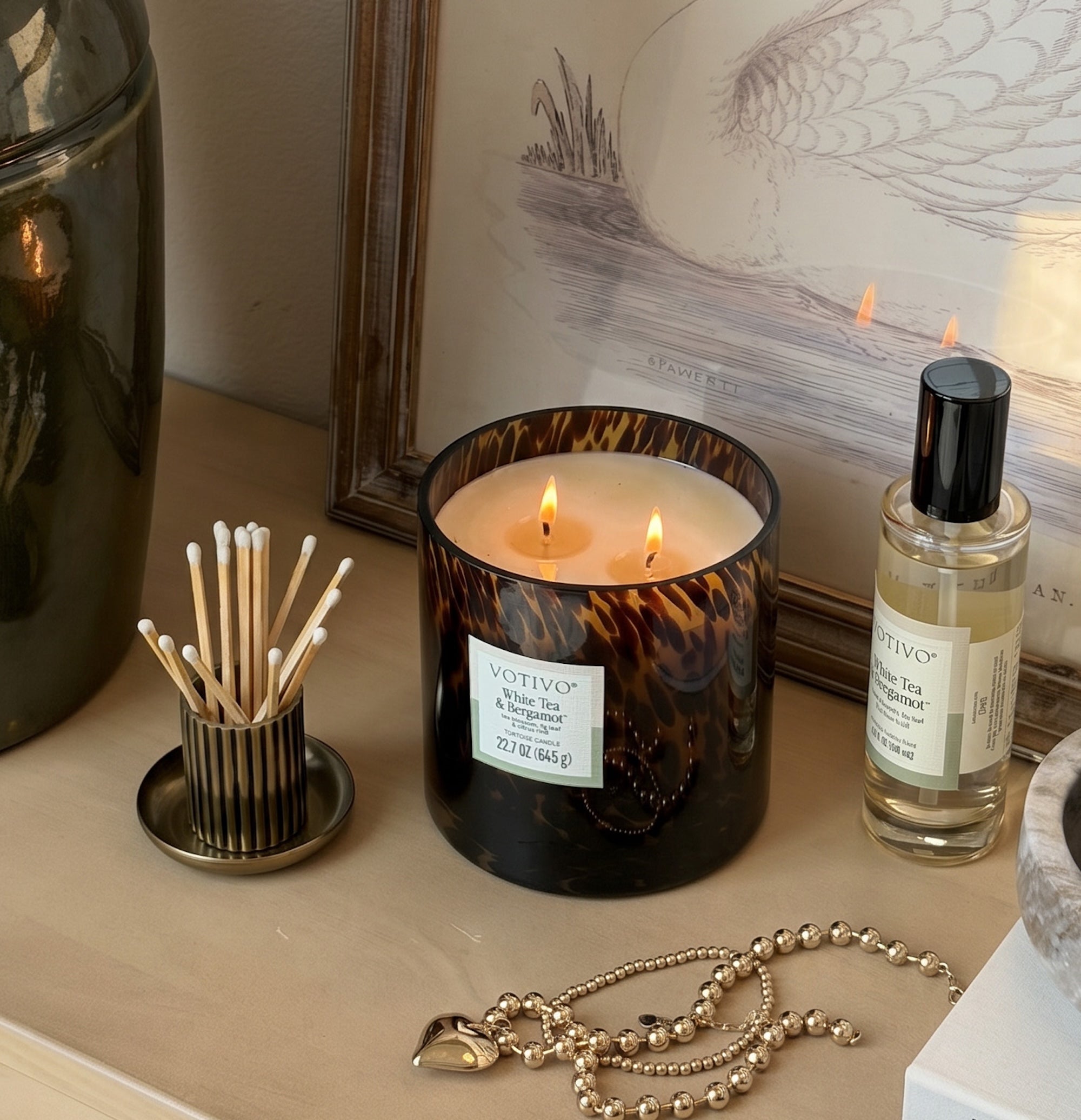 A Votivo White Tea & Bergamot Tortoise Candle rests on a table beside a room spray, match container, and gold heart charm necklace, with framed art in the background. The candle is housed in brown marbled glass.