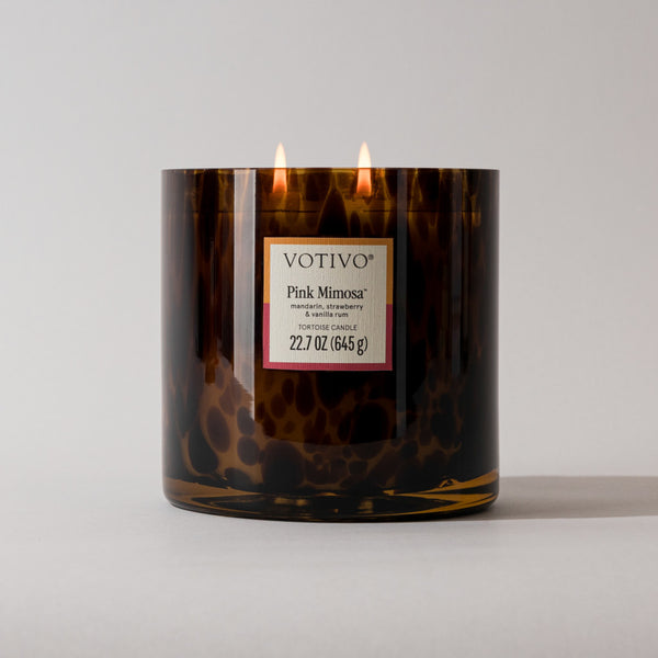 A brown, speckled glass soy blend candle with two lit wicks sits against a white background. Its label reads Votivo Pink Mimosa Tortoise Candle. The candle weighs 22.7 oz (645 g).