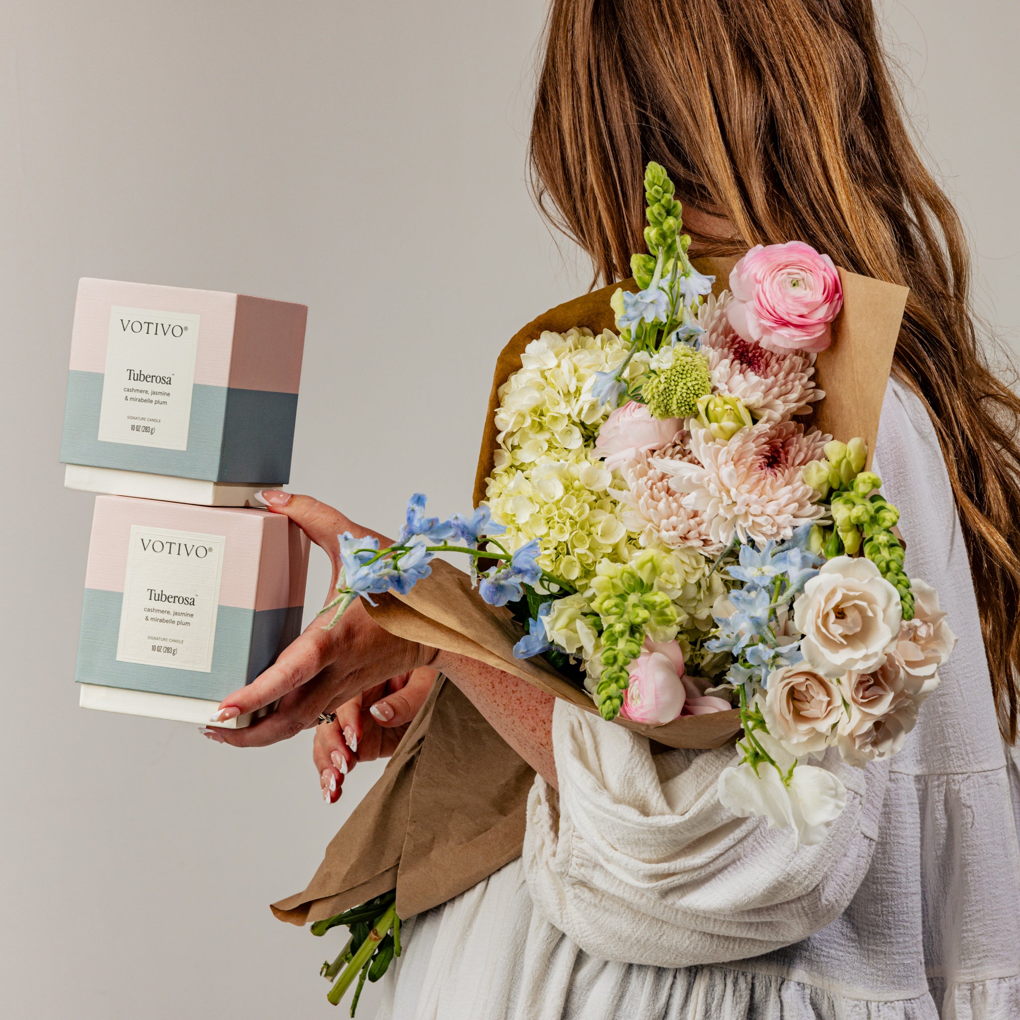 A woman in a white shirt holds a bouquet of pastel flowers in one arm and two Votivo Tuberosa 10 oz. Signature Candles in the other hand against a plain background.