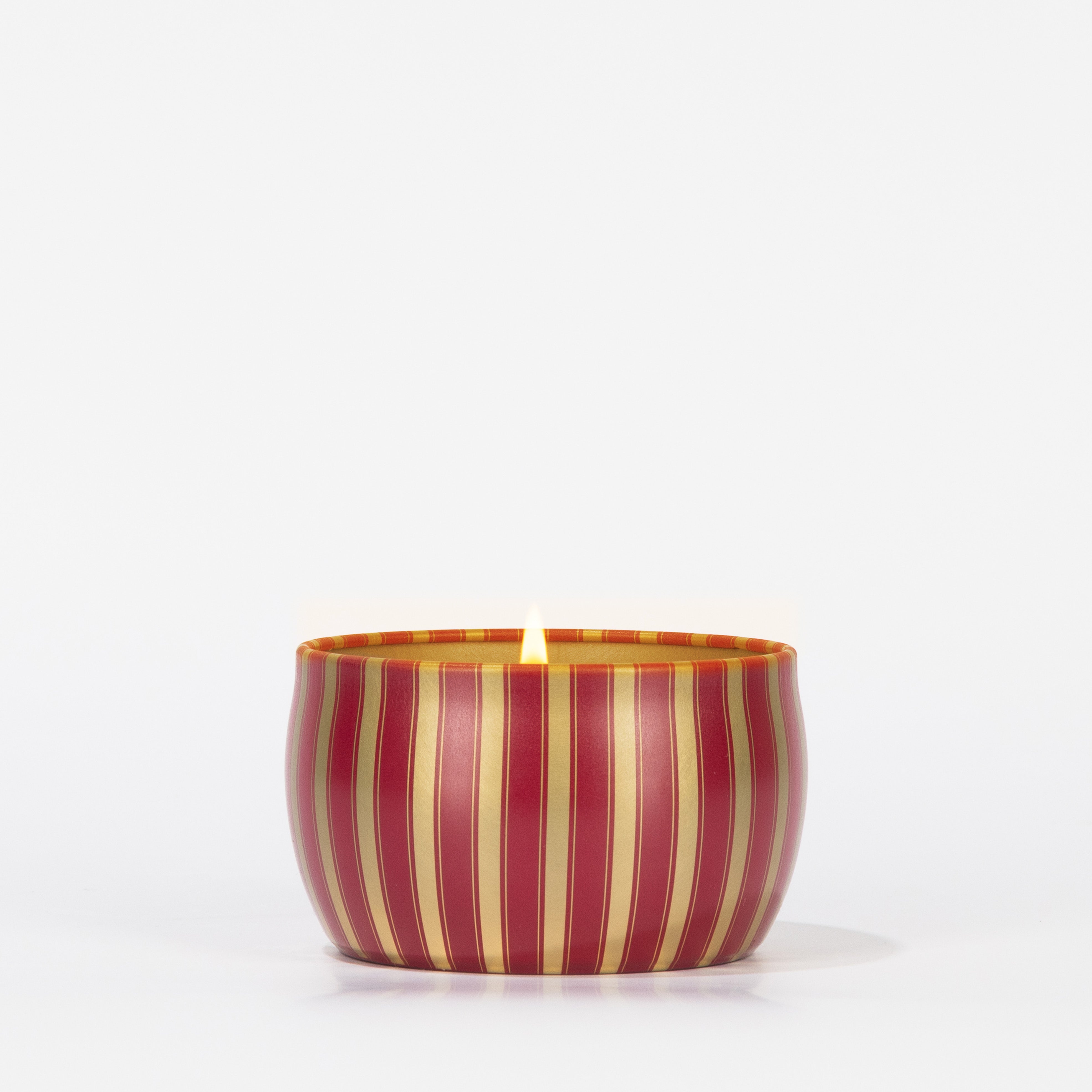 A Votivo Velvet Rouge Holiday Tin Candle, made from soy blend wax, sits in a round tin with vertical red and gold stripes on a white background.
