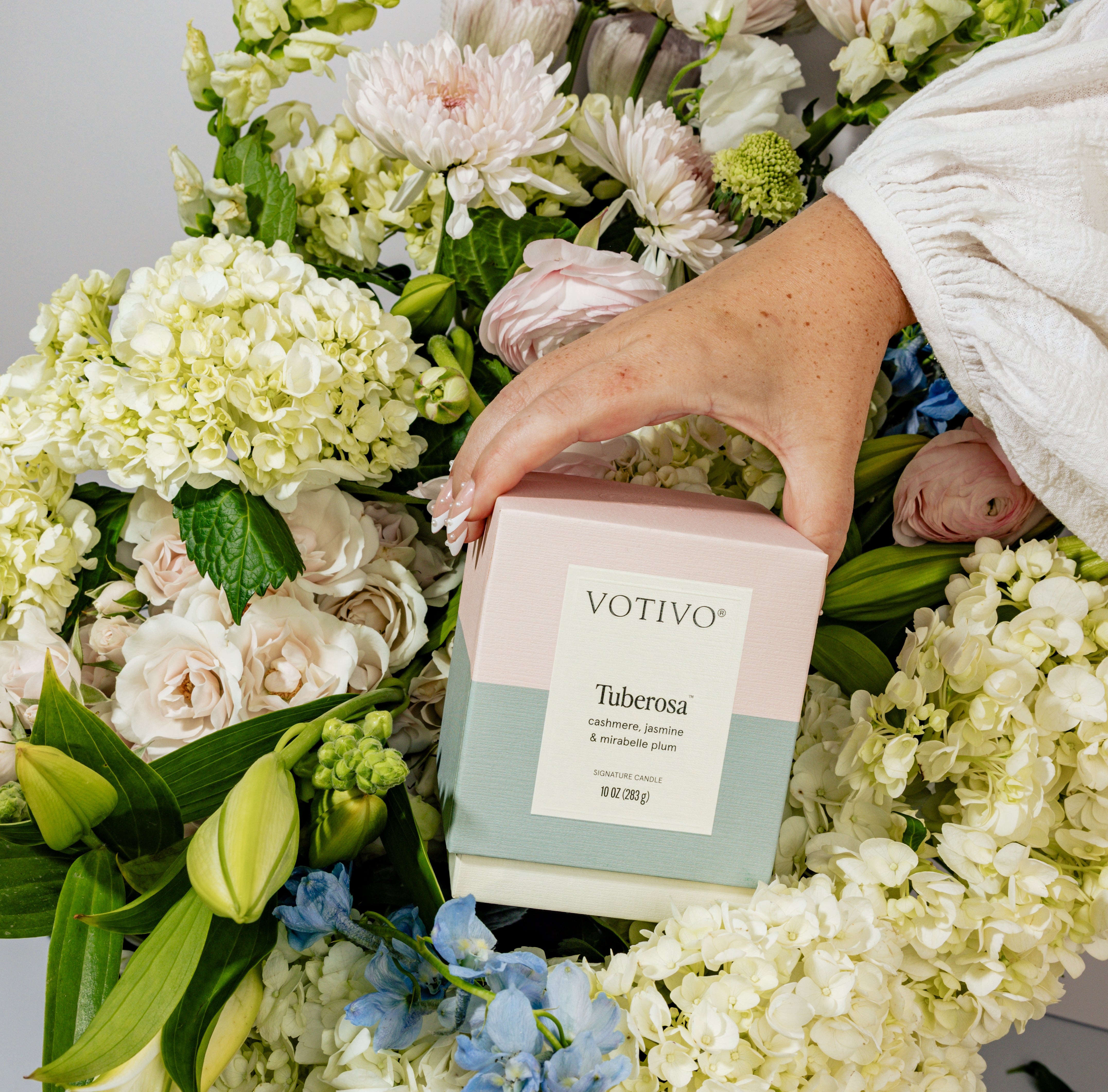 A hand in a white sleeve holds the Votivo Tuberosa 10 oz. Signature Candle box, made with soy blend wax, surrounded by lush white, pink, and green flowers.