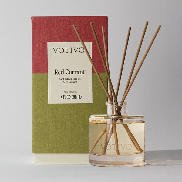 A Votivo Red Currant Reed Diffuser features fiber reeds in a clear glass bottle, displayed with its red and olive green box, and highlights scent notes of tart citrus, cassis, and geranium.