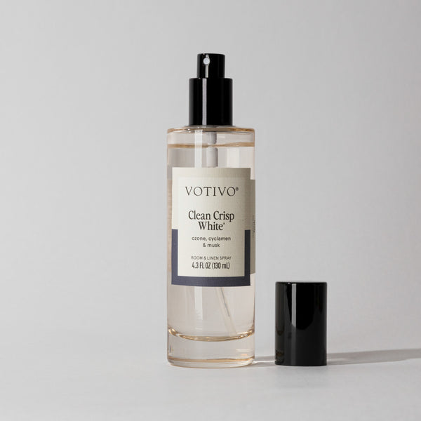 A bottle of Votivo Clean Crisp White Room & Linen Spray, featuring a black cap, sits on a simple light background. The label emphasizes this fresh scent, ideal for brightening any space.