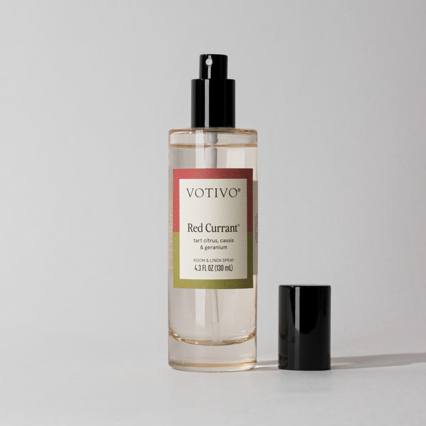 A Votivo Red Currant Room & Linen Spray with a black cap rests on a light gray background, its label emphasizing a fruity scent blending tart citrus, cassis, and geranium.