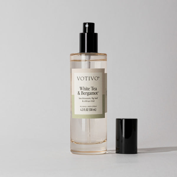 A clear Votivo White Tea & Bergamot Room & Linen Spray bottle with a black nozzle and fabric-safe black cap sits on a plain light gray background.