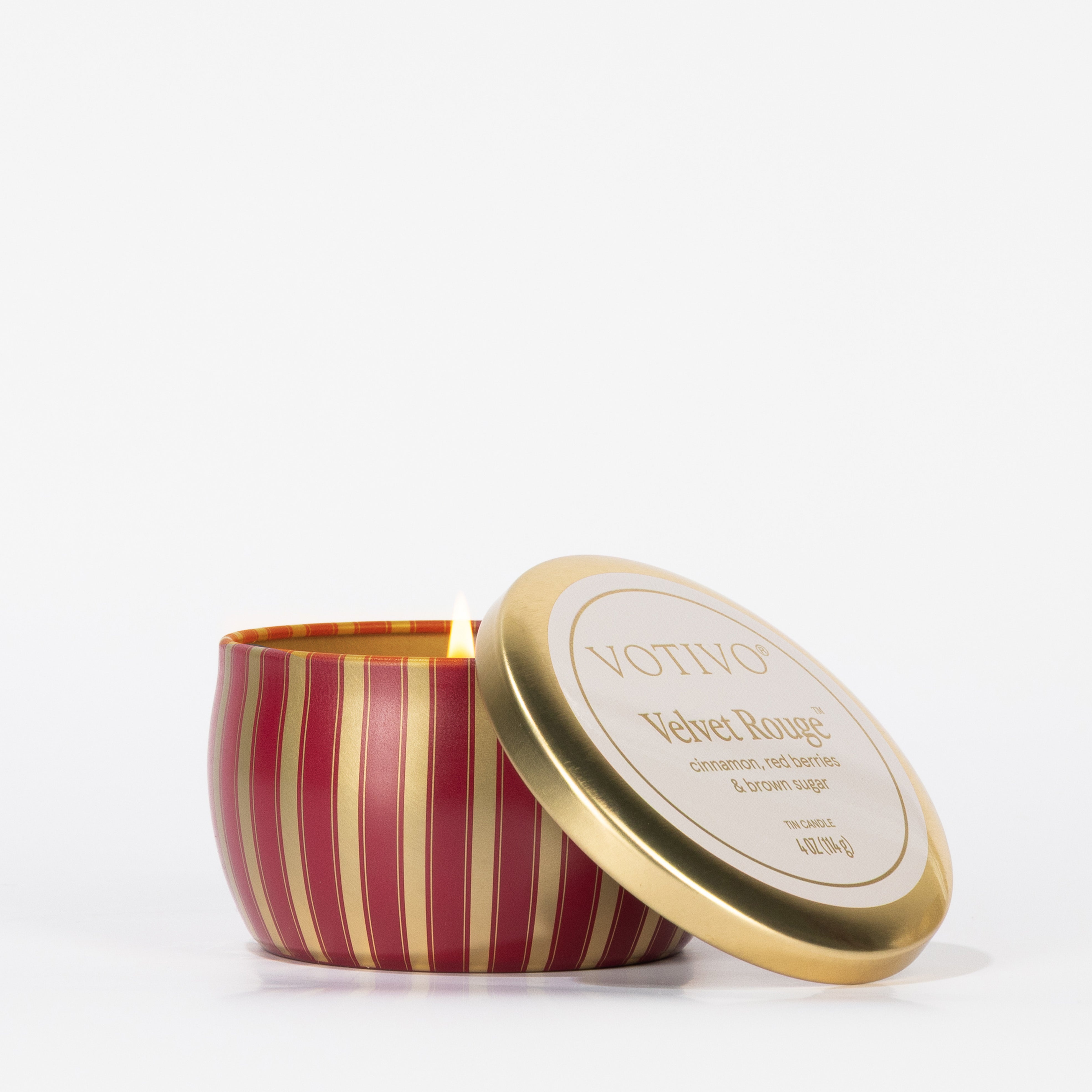 A lit Votivo Velvet Rouge Holiday Tin Candle in a red and gold striped tin sits on a white background, its labeled lid placed beside it.