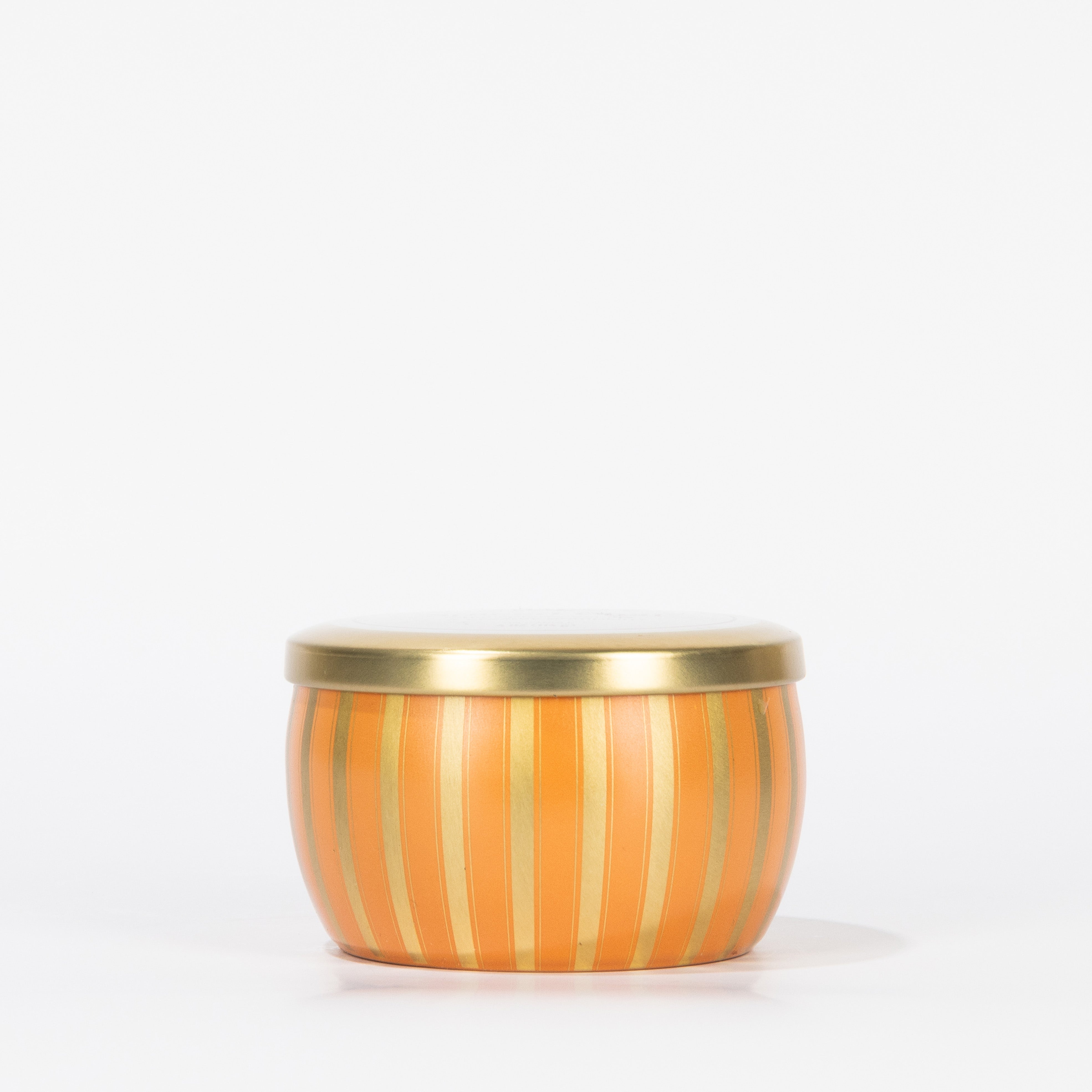 The Votivo Spiced Chai Holiday Tin Candle is a small, round soy blend candle with vertical orange and gold stripes and a flat metallic gold lid, shown against a plain white background.