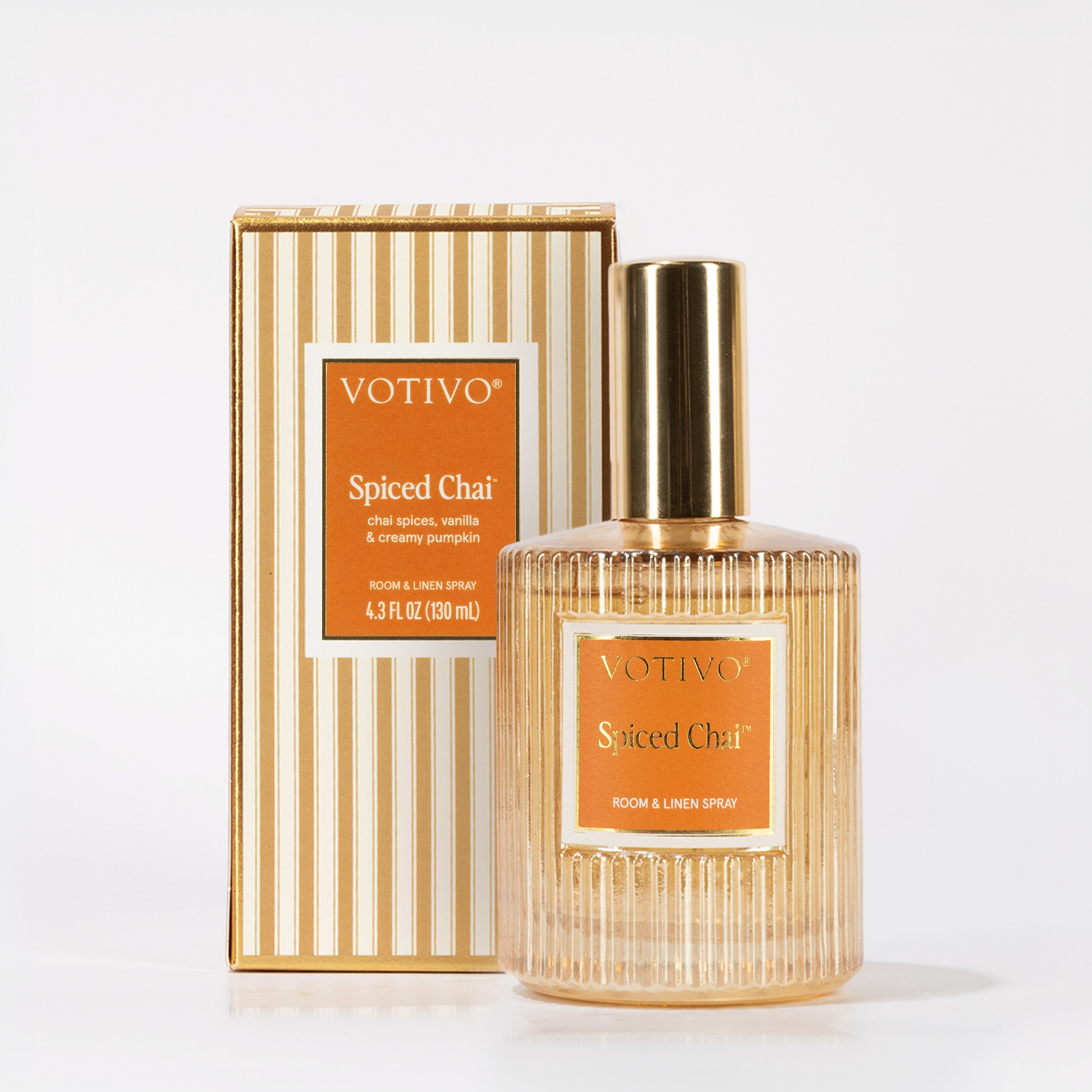 A Votivo Spiced Chai Room & Linen Spray with a gold cap is displayed before its matching striped box. The orange label features white text, and the spray is fabric safe for versatile holiday freshness.
