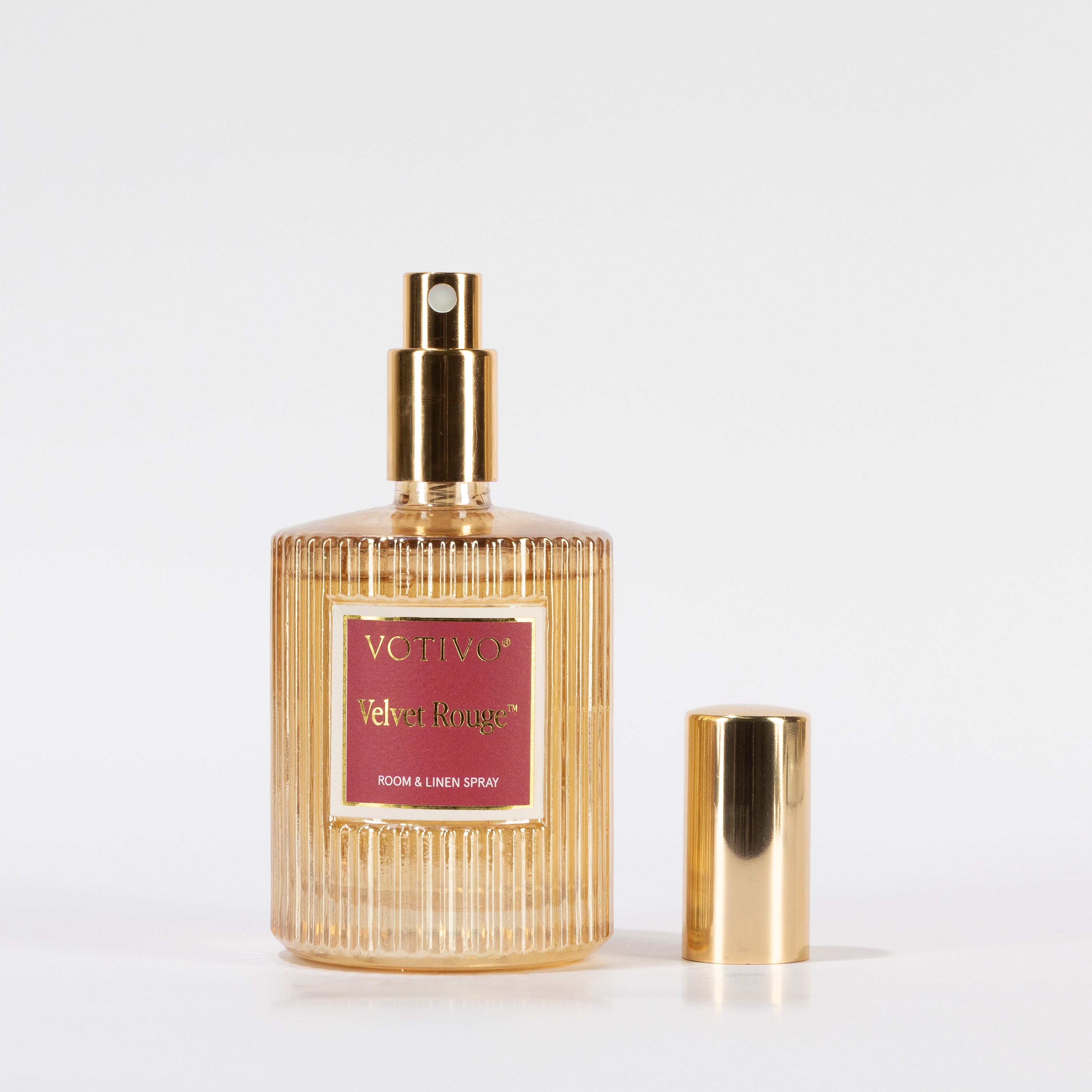 A gold, ribbed glass bottle of Votivo Velvet Rouge Room & Linen Spray with a gold spray nozzle and matching cap sits beside it on a plain white background.