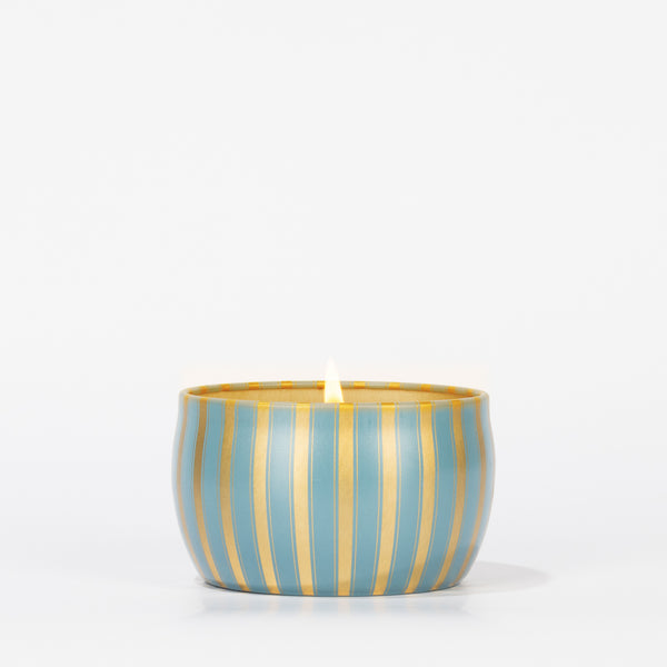 The Votivo Icy Blue Pine Holiday Tin Candle, in a round blue and gold striped container filled with soy blend wax, is displayed centered on a plain white background.