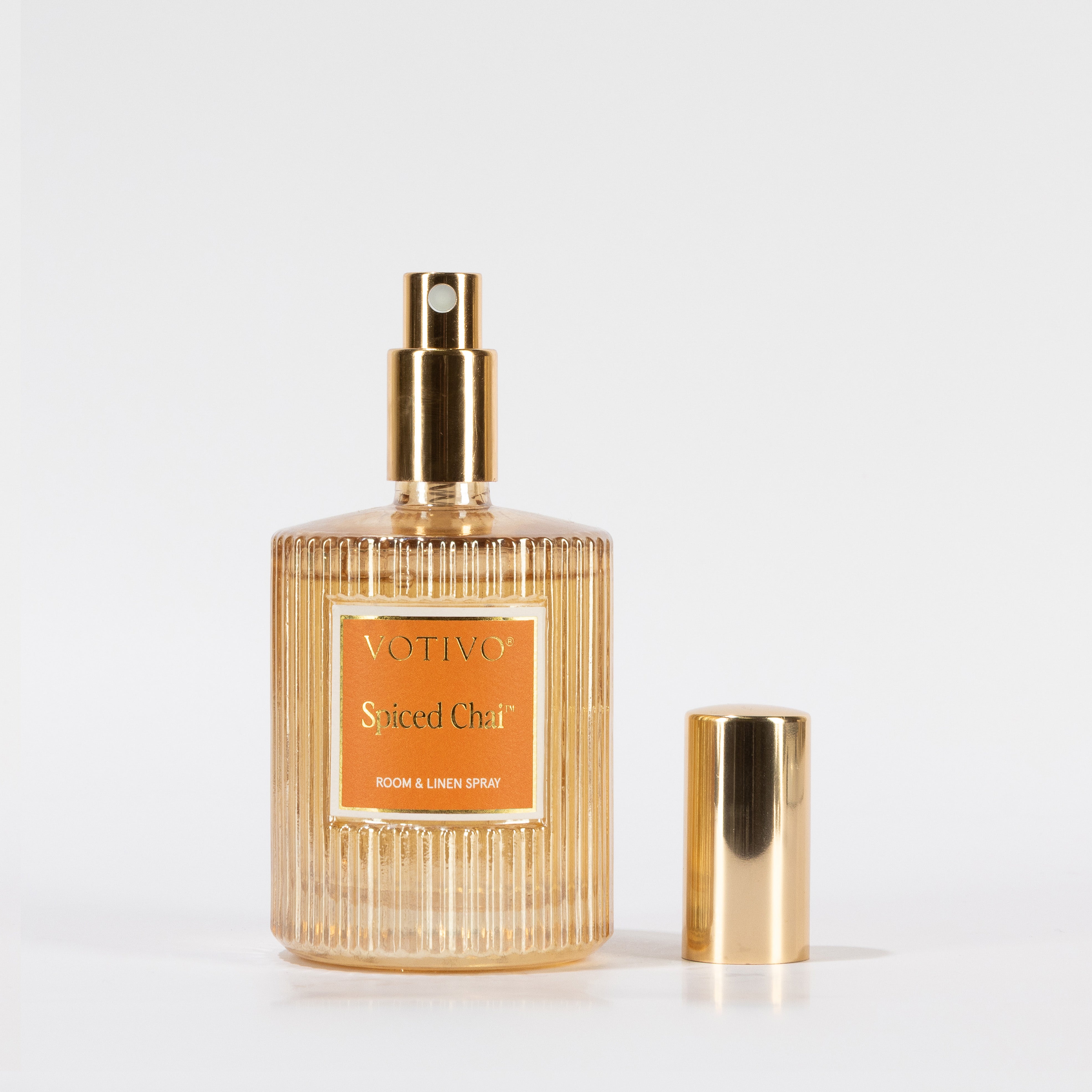 A gold and clear ribbed glass bottle labeled “Votivo Spiced Chai Room & Linen Spray” stands upright with its gold cap beside it on a white background, adding a warm, inviting scent to any room or fabric.