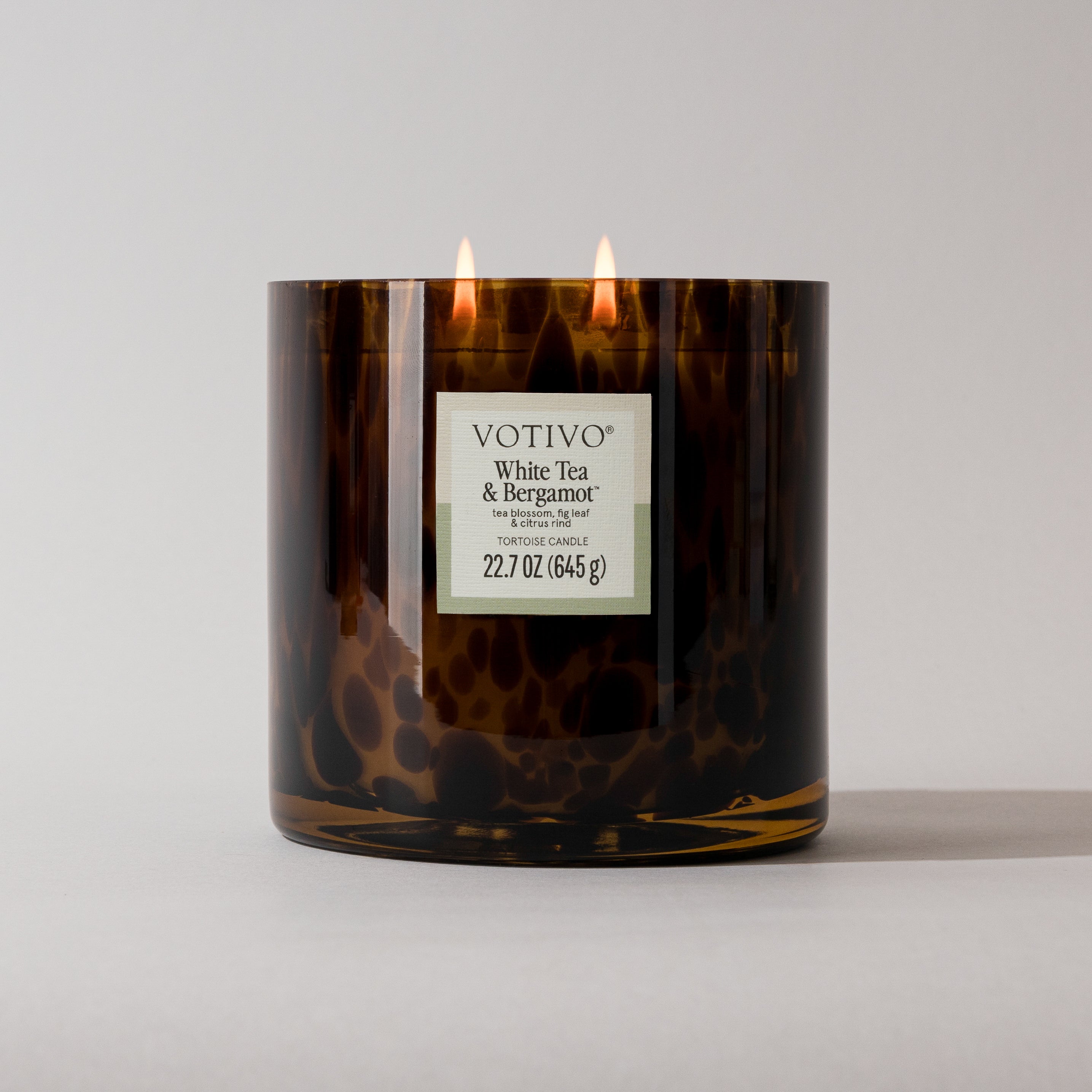 A Votivo White Tea & Bergamot Tortoise Candle featuring a brown, speckled design and two lit wicks rests on a light gray surface with a plain gray background.