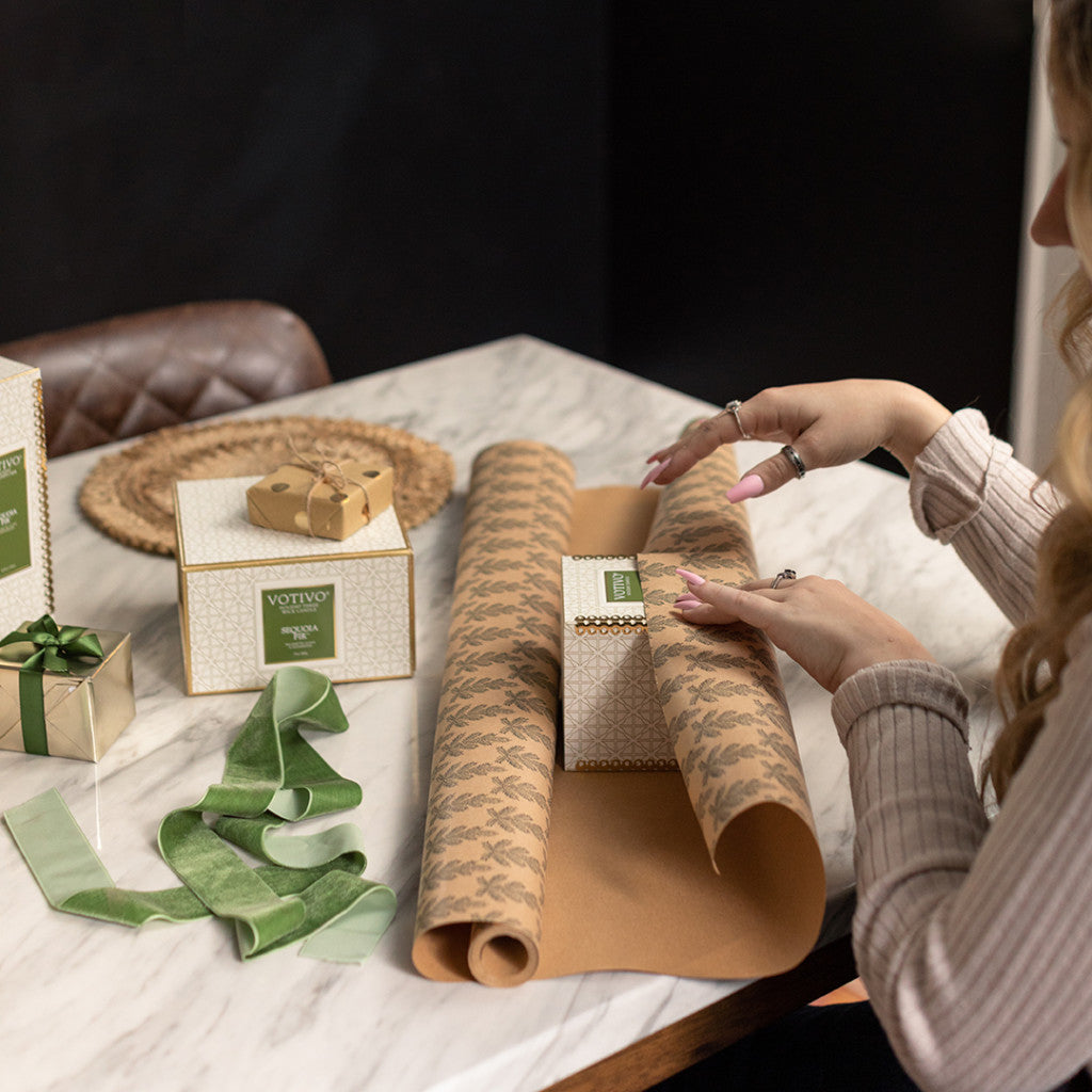 A person wraps a Votivo gift box in brown patterned paper on a marble table with scissors, green ribbon, and other gifts—perfect for gifting the Sequoia Fir Holiday 3 Wick Candle by Votivo.
