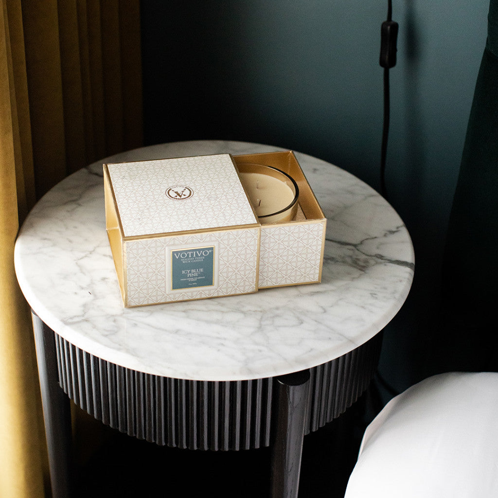A Votivo Icy Blue Pine Holiday 3 Wick Candle in an open, patterned box rests on a round white marble side table near a bed, with a curtain and wall-mounted cord in the background. Made with premium soy blend wax.